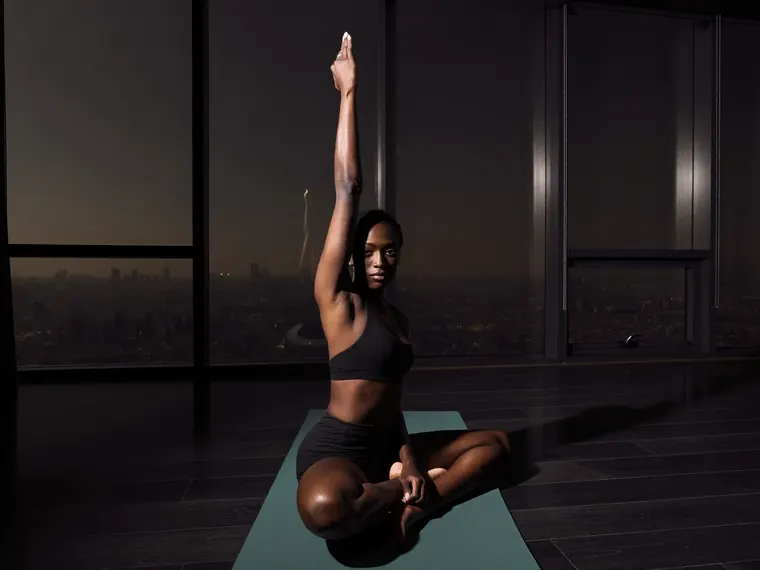 A woman sits cross-legged on a green mat, one arm raised, a city at dusk behind her