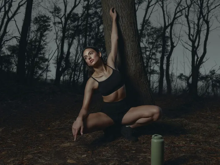 A woman stretches against a tree in a dark forest, a green bottle at her feet