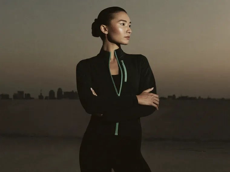 A woman in a black jacket with a green zip stands with arms folded against a city skyline at dusk