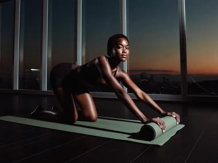 A woman rolls up a green yoga mat at sunset in front of floor-to-ceiling windows