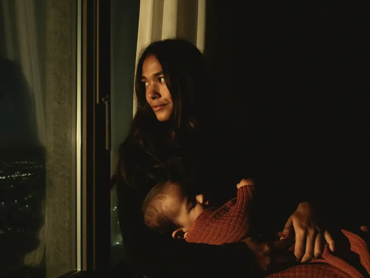 A mother holds her sleeping baby, wrapped in a terracotta knit, by a window at night with city lights below