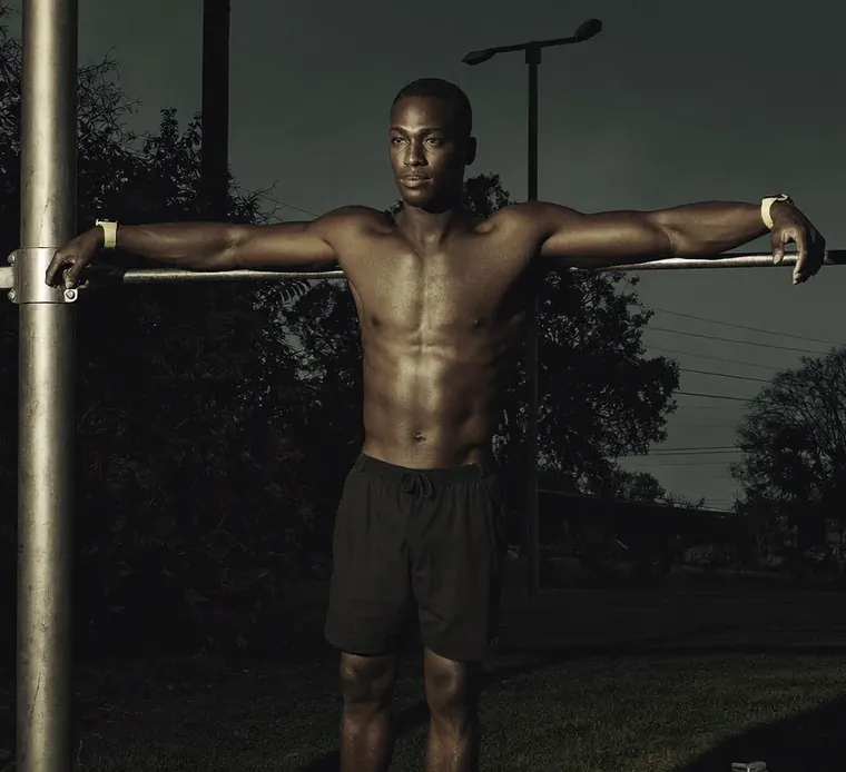 A man rests his arms along the bars of an outdoor gym at night, trees and streetlights behind him