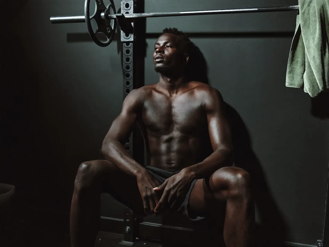 A man rests against a squat rack in a dark home gym, a green towel hanging beside him