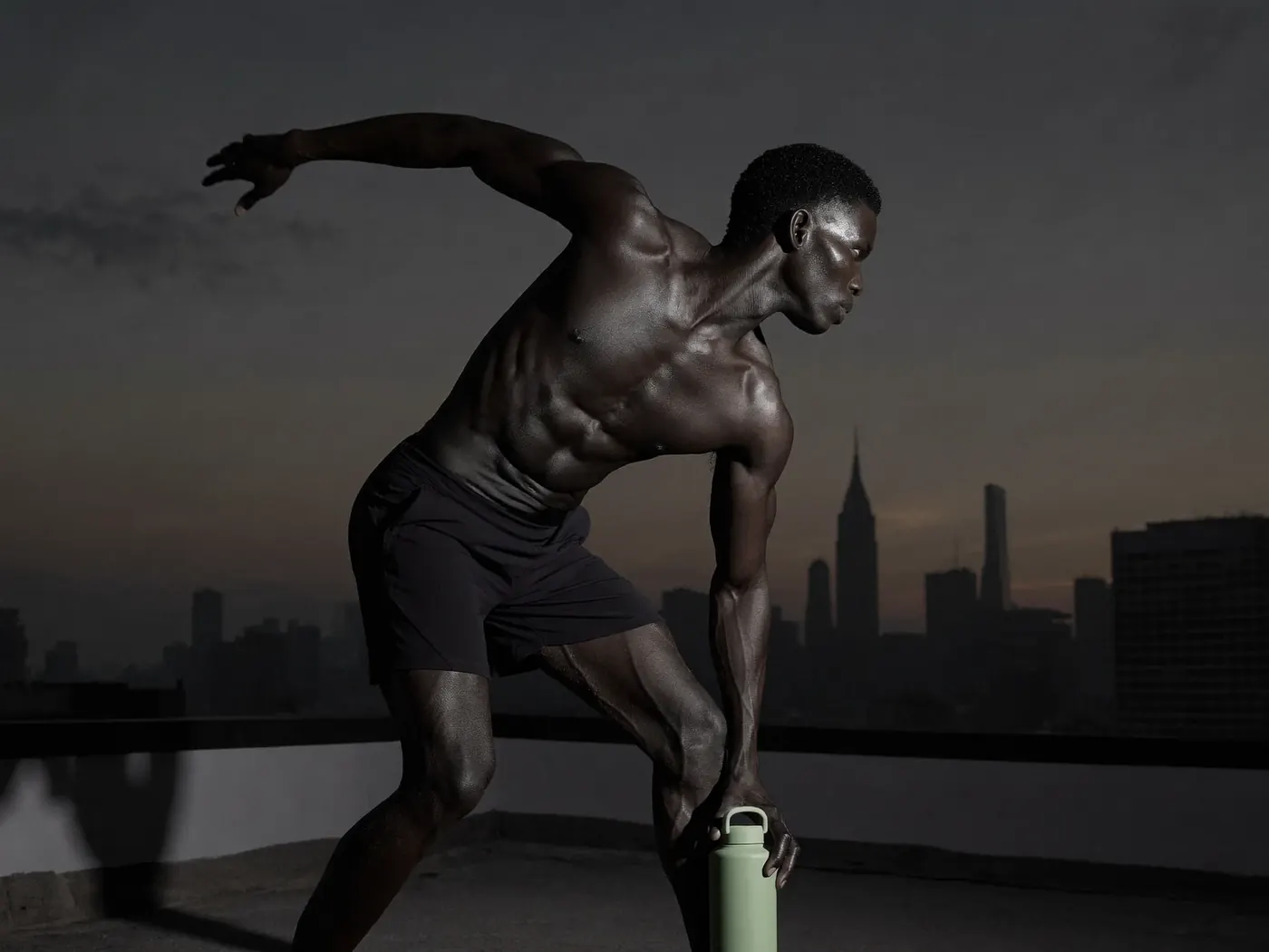 A man stretches on a rooftop at dusk, one hand on a green water bottle, a city skyline behind him