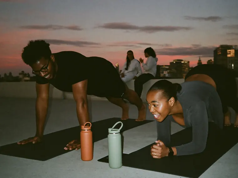 Two friends laugh through a plank on a rooftop at dusk, terracotta and sage bottles between their mats, two more friends on the ledge behind them